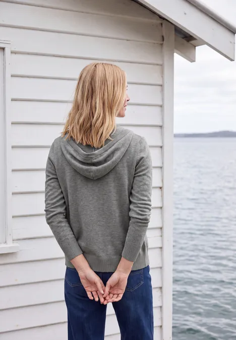 Langarm Cardigan mit Kapuze sporty grey melange Frau im grauen Kapuzenpullover steht mit verschränkten Händen vor weißer Holzhütte am Meer.