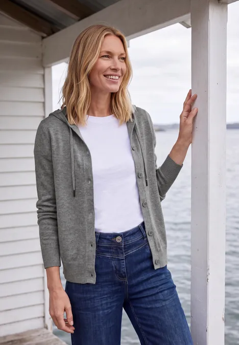 Langarm Cardigan mit Kapuze sporty grey melange Lächelnde Frau mit grauer Kapuzenjacke, weißem T-Shirt und blauen Jeans lehnt an einem weißen Holzhaus am Meer