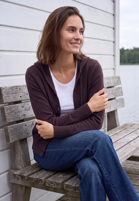 Langarm Cardigan mit Kapuze eggplant red melange Eine lächelnde Frau sitzt im Pullover und Jeans auf einer Holzbank vor einer weißen Wand am Wasser.