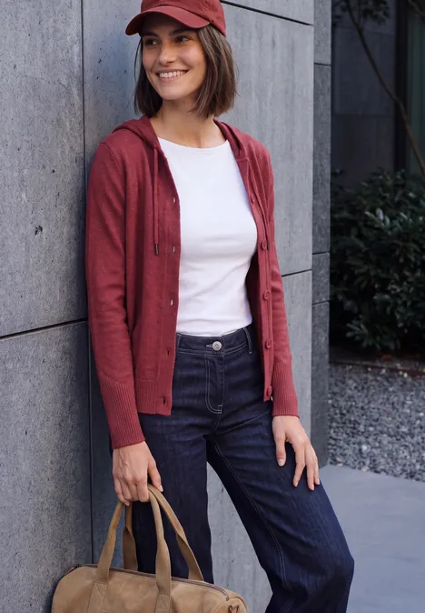 Frau an Wand, lächelnd mit roter Mütze & Strickjacke, weißem T-Shirt, dunkler Hose und beiger Tasche.