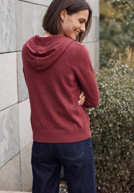 Frau mit bordeauxfarbener Kapuzenjacke und Jeans lehnt lächelnd an einer Steinmauer und einem Busch.