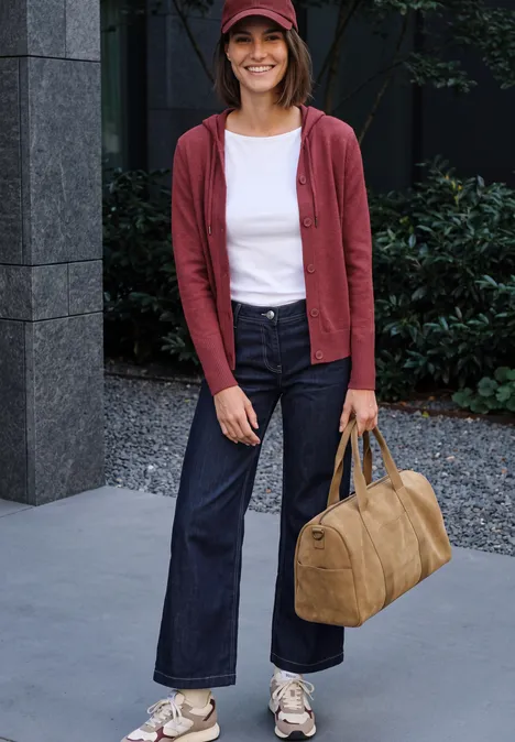 Junge Frau in roter Jacke, Jeans und roter Mütze trägt eine Tasche, lächelt vor einem Gebäude.