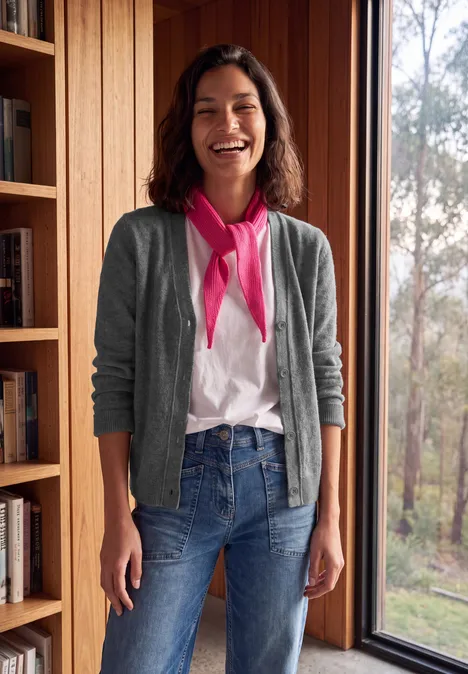Een lachende vrouw in jeans, wit t-shirt en grijze cardigan staat nonchalant voor een raam in een houten interieur.