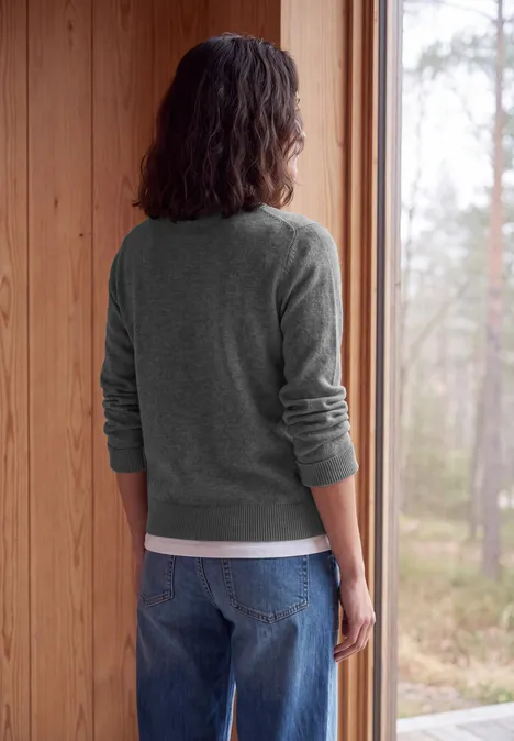Een persoon in een grijze trui en spijkerbroek staat in een houten kamer en kijkt naar het bos buiten.