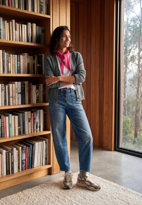 Een vrouw staat nonchalant voor een houten boekenrek, in jeans en vest, en kijkt uit het raam.