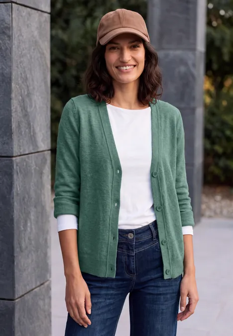 Glimlachende vrouw in groene cardigan en spijkerbroek leunt buiten tegen een stenen muur.
