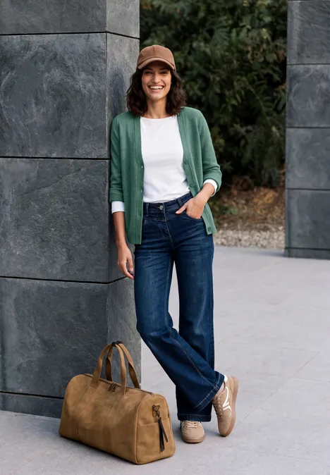 Vrouw in jeans, wit t-shirt en groene cardigan met hoed leunt tegen een muur, tas op de grond.