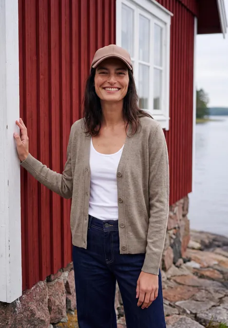 Een glimlachende vrouw in jeans, vest en pet voor een rood houten huis aan het water.