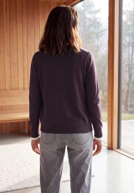Frau in lila Pullover und Jeans steht in einem hellen Holzraum vor einem großen Fenster.