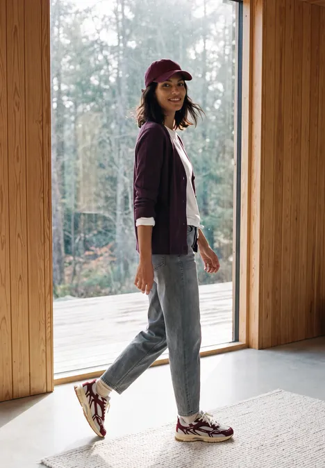 Frau in Jeans, Sneakern, Hoodie und Cap steht in einem hellen Holzhaus vor einer großen Glasfront.