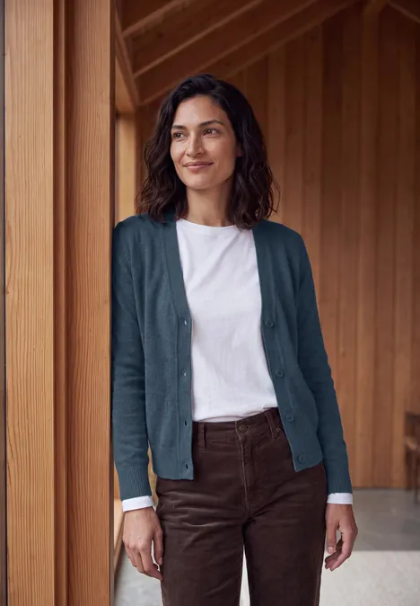 Eine Frau mit weißem T-Shirt, blauem Cardigan und brauner Hose vor Holzwand.