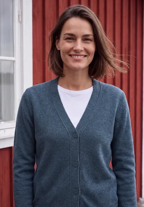 Eine Frau mit kurzem braunem Haar lächelt in einem grauen Cardigan vor einem roten Holzhaus.
