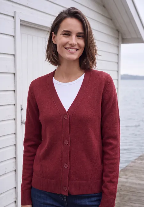 Vrouw met rode vest en wit shirt lacht op een steiger voor een wit huis aan het water.