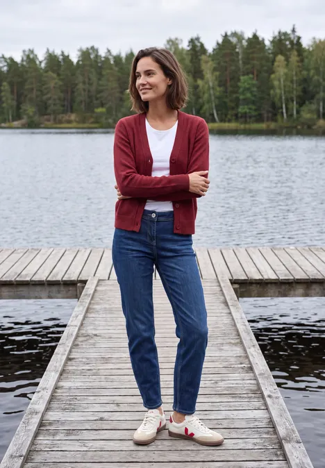 Een vrouw in een rode cardigan staat glimlachend op een houten steiger bij een meer, met bomen op de achtergrond.