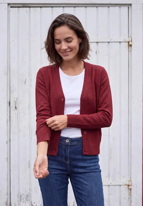 Een vrouw in een rood vest, wit shirt en spijkerbroek glimlacht voor een witte houten deur.