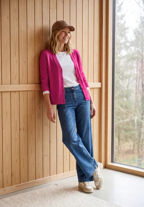 Vrouw leunt ontspannen tegen houten wand bij raam, draagt roze blazer, wit shirt, spijkerbroek, sneakers en pet.