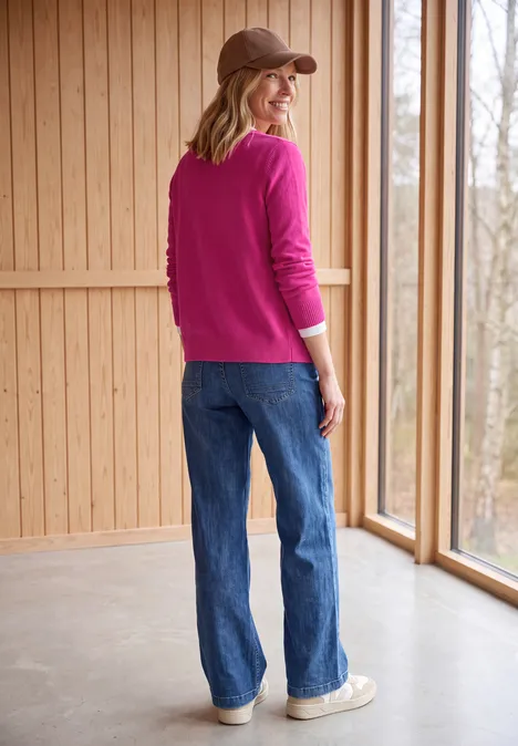 Vrouw met pet, fuchsia trui en jeans staat bij een raam in een lichte houten ruimte en kijkt over haar schouder.