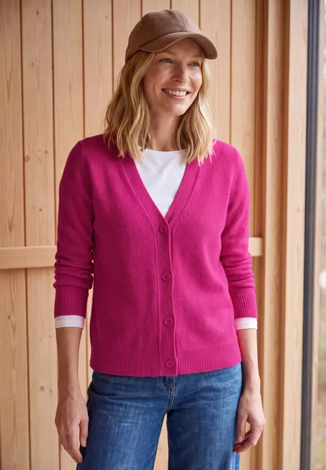 Lachende vrouw in roze vest, wit shirt, jeans en bruin petje voor een houten muur.