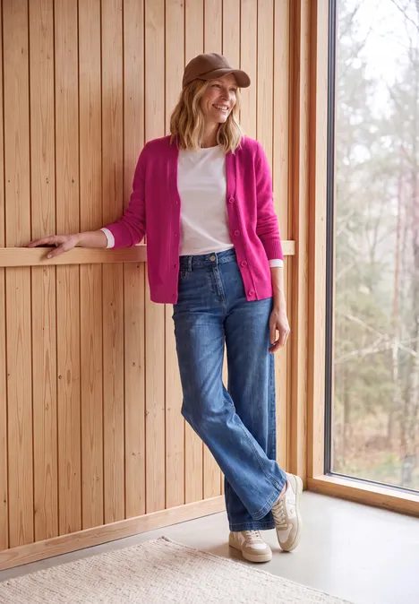 Vrouw lacht geleund tegen houten wand, draagt roze vest, wit shirt, jeans en hoed.