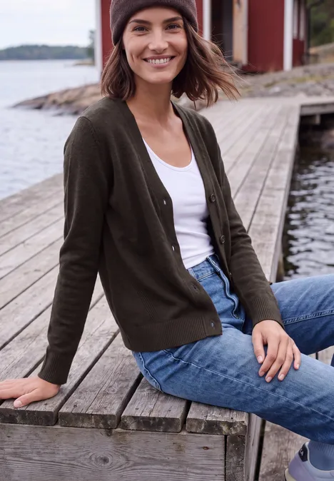 Junge Frau sitzt lächelnd auf einem Holzsteg am Wasser in Strickjacke, Jeans und Mütze.