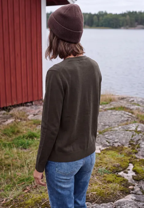 Person in Strickpullover, Jeans und Mütze steht am Felsufer vor einem roten Gebäude mit Blick aufs Wasser.