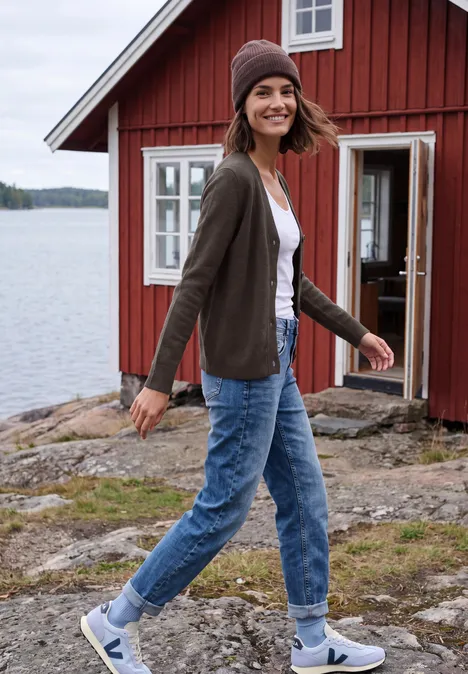 Frau in Strickjacke, Jeans und Mütze geht an einem roten Holzhaus am See spazieren.