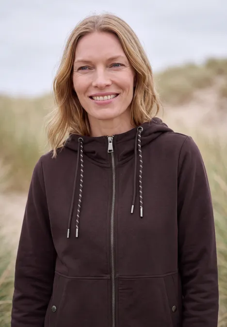 Sweatjacke mit Zipper und Kapuze berry red Eine lächelnde Frau in brauner Kapuzenjacke steht vor einer Dünenlandschaft.