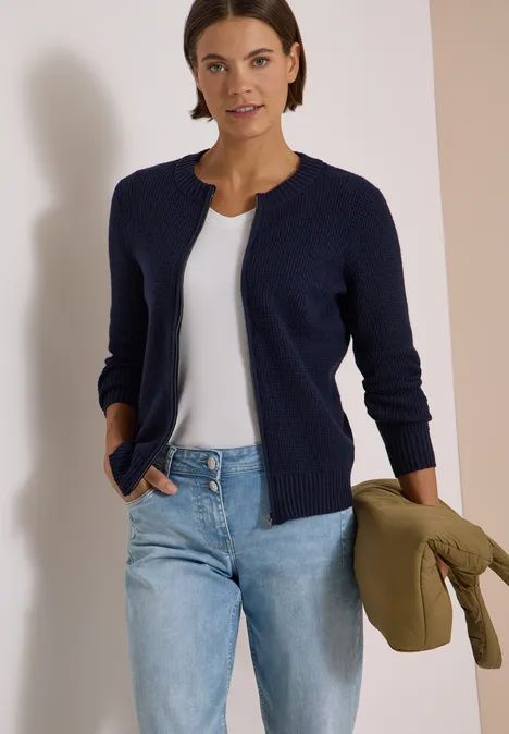 Frau trägt dunkelblauen Cardigan, weiße Bluse, Jeans, hält olivgrünen Mantel lässig unter dem Arm.