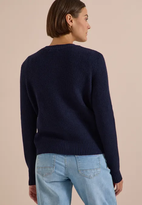 Frau mit Kurzhaarschnitt trägt von hinten einen dunkelblauen Pullover und hellblaue Jeans vor hellem Hintergrund.