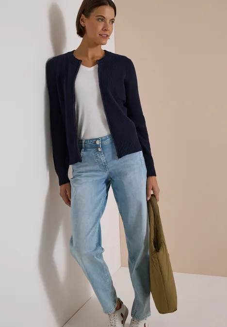 Frau in weißem Shirt, hellblauen Jeans, navy Cardigan und grüner Tasche vor zweifarbigem Hintergrund.