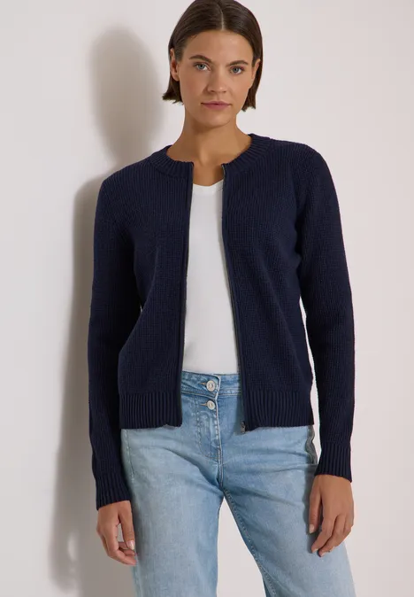 Frau in dunkelblauer Strickjacke, weißem Top und Jeans vor hellem Hintergrund.