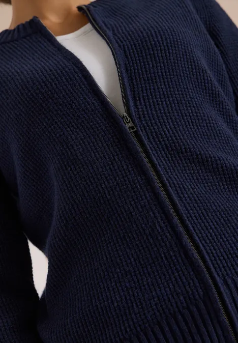 Dunkelblauer, grob gestrickter Cardigan mit Reißverschluss, offen sichtbar ein weißes Top darunter.