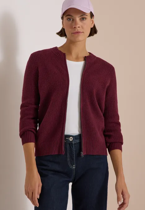 Frau in Jeans, weißem Shirt, burgunderrotem Cardigan und Cap vor hellem Hintergrund.