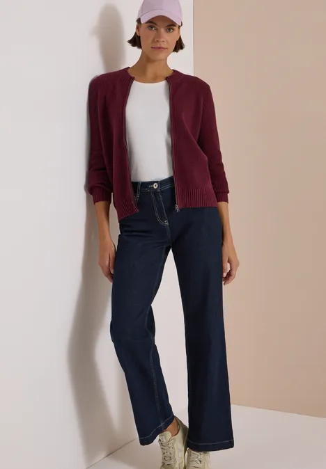 Frau in weißem Shirt, bordeauxfarbenem Blazer, weiter Jeanshose und rosa Cap vor hellem Hintergrund.