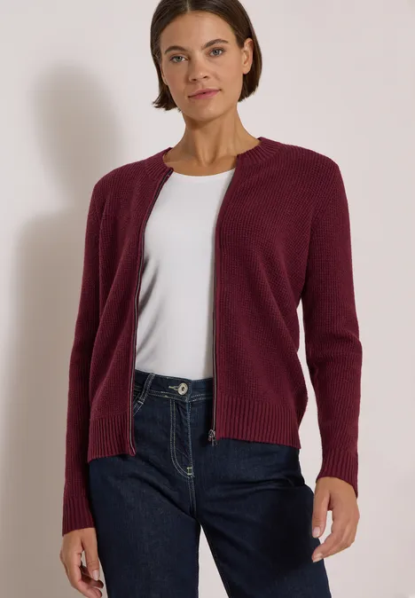 Eine Frau trägt einen bordeauxroten Cardigan, weißes Shirt und blaue Jeans.