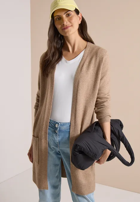 Langer Cardigan in offener Passform creamy mocha melange Frau lehnt an Wand, trägt beige Strickjacke, weißes Top, Jeans und gelbe Cap, hält schwarze Tasche.