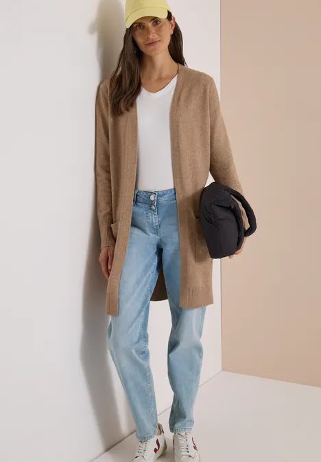 Langer Cardigan in offener Passform creamy mocha melange Frau trägt lange beige Strickjacke, weißes V-Shirt, helle Jeans, weiße Sneaker, hält Mütze und Jacke