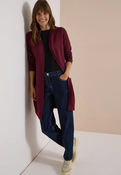 Langer Cardigan in offener Passform red wine Eine Frau lehnt lächelnd an der Wand, trägt einen burgunderfarbenen Cardigan, Pullover, Jeans und helle Schuhe.