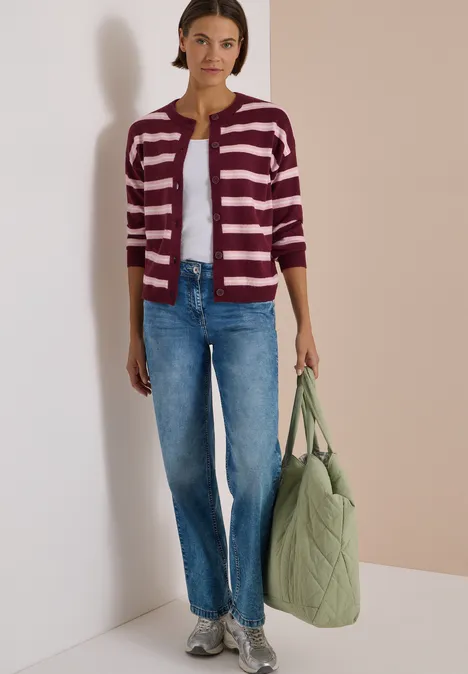 Frau trägt gestreifte Strickjacke, blaue Jeans, Sneaker und hält eine grüne, gesteppte Tasche.