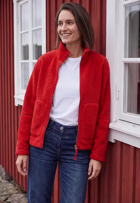Lächelnde Frau in roter Fleecejacke, weißem Shirt und Jeans lehnt vor einem roten Haus mit weißen Fensterrahmen.
