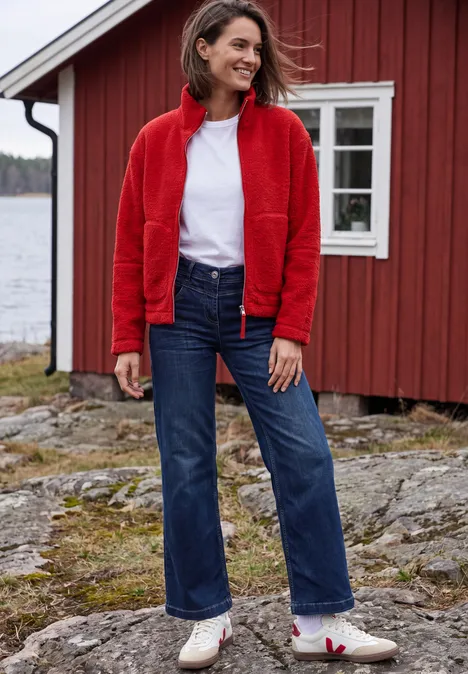 Frau in roter Jacke, weißem Shirt und Jeans steht vor einem roten Holzhaus am See auf felsigem Untergrund.