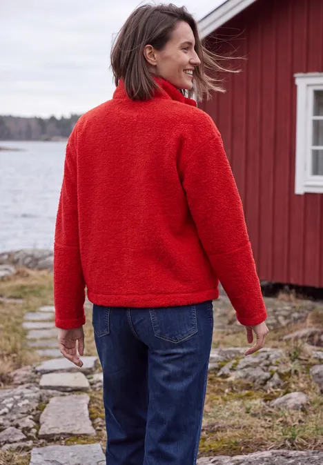 Frau in roter Fleecejacke und Jeans steht am Seeufer vor einem roten Haus.