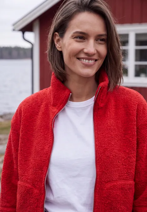 Eine lächelnde Frau in roter Fleecejacke und weißem T-Shirt steht vor einem roten Haus am See.