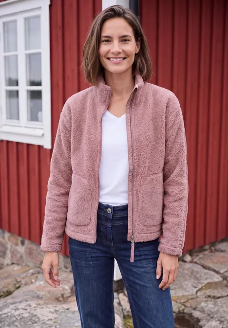 Eine lächelnde Frau in rosa Fleecejacke, weißem T-Shirt und Jeans vor einem roten Holzhaus.