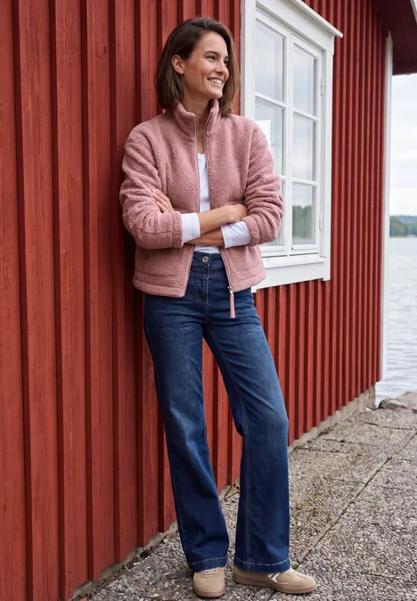 Eine lächelnde Frau in rosa Jacke und Jeans lehnt entspannt an einem roten Holzhaus am Wasser.