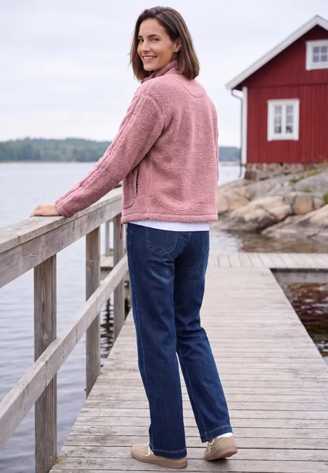 Frau in rosa Jacke und Jeans lehnt an Geländer auf einem Holzsteg an einem roten Haus am Wasser.