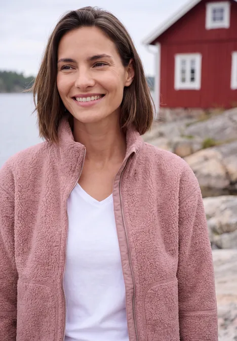 Eine lächelnde Frau in rosa Fleecejacke vor einem roten Haus am Wasser.