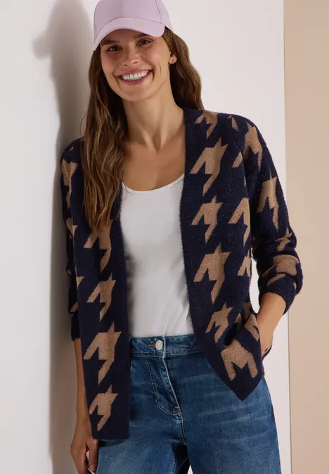 Lächelnde Frau in Hahnentritt-Strickjacke, weißem Top, Jeans und Cap