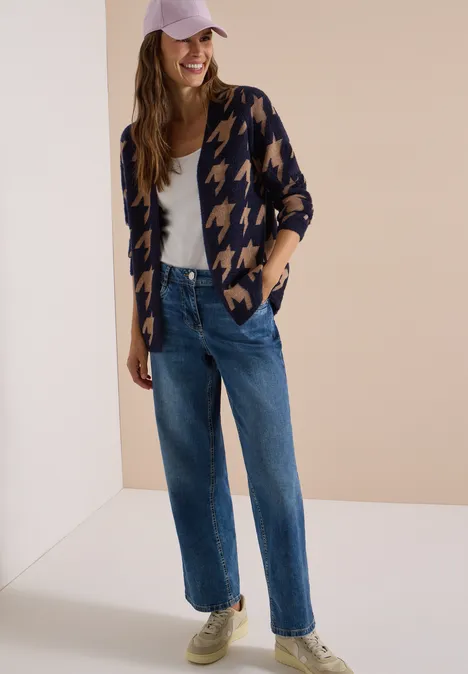 Frau in geraden Jeans, weißem Top, Hahnentritt-Strickjacke und Cap, lächelnd vor neutralem Hintergrund.