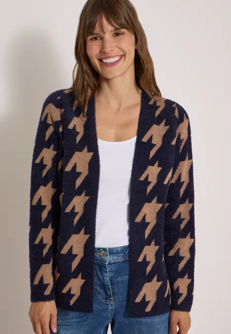 Frau mit offenem Cardigan im Hahnentrittmuster (Marine/Beige), weißem Top und Jeans, lächelt in die Kamera.